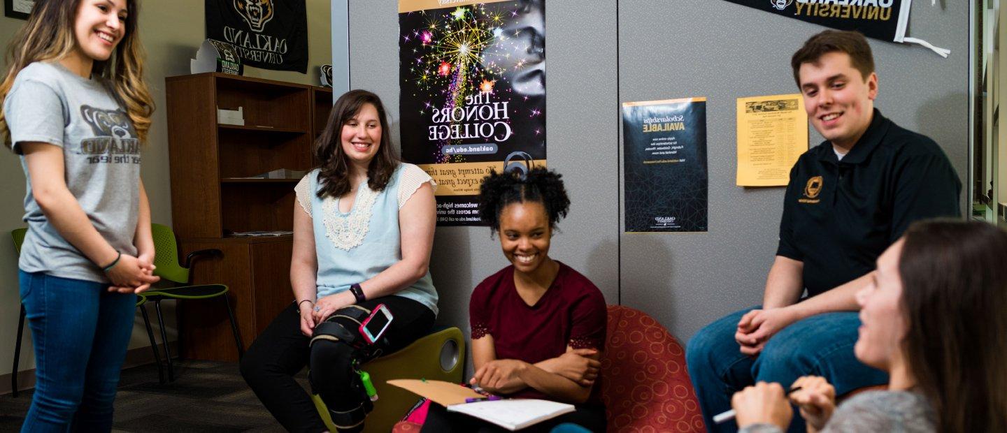 A group of young people sits in an office, talking and laughing. An Honors College poster can be seen in the background.